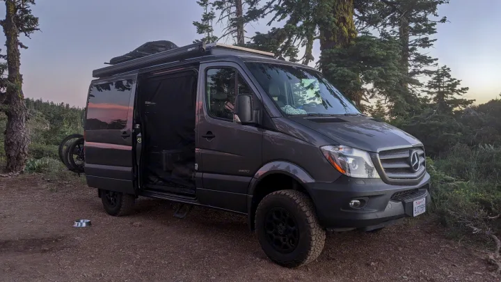 A custom dark gray Mercedes Sprinter van, perfect for an rv rental, parked on a dirt trail in a forest with an open side door.. Mercedes-Benz Sprinter 2016