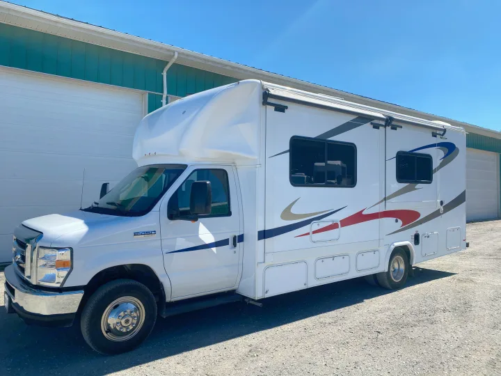 White Class C RV rental with blue and red accents, parked outdoors in front of a green building.. Ford E450 2013