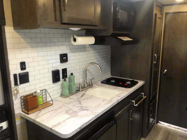 Modern RV rental kitchen featuring a subway tile backsplash, marble-look counter, sink, and cooktop. Compact & functional!. Jayco Jay Flight 2021