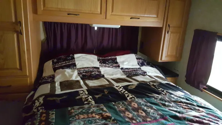 An RV rental's cozy bedroom, showcasing a patterned quilt, wooden overhead storage, and a side window.. Ford Majestic 2007