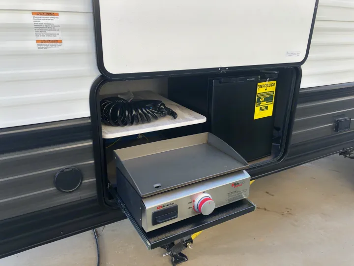 This rv rental's outdoor kitchen features a pull-out griddle, mini-fridge, and hose, with visible safety warnings.. Forest River Wildwood X-Lite 2020