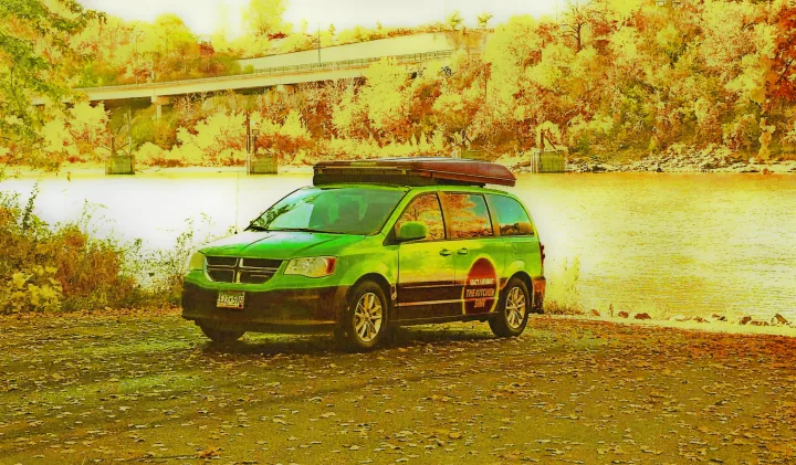 A vibrant green minivan RV rental with a rooftop box is parked by a river, surrounded by autumn trees.. Dodge Grand Caravan 2014