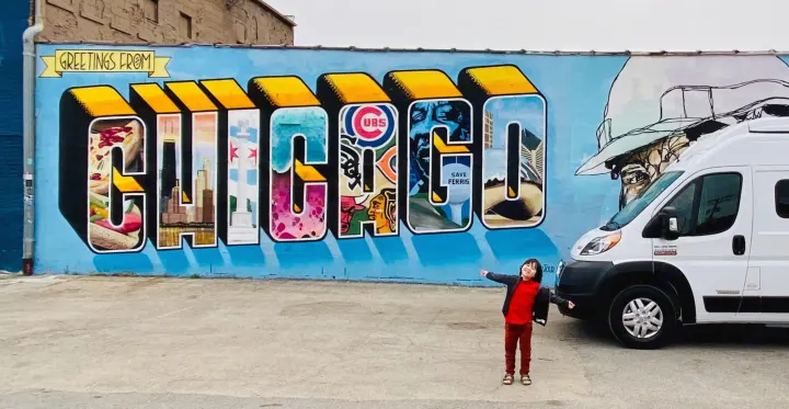 A child poses excitedly before a colorful 'Greetings from Chicago' mural. A white RV rental is parked nearby for their family travels.. Winnebago Solis 59PX 2021