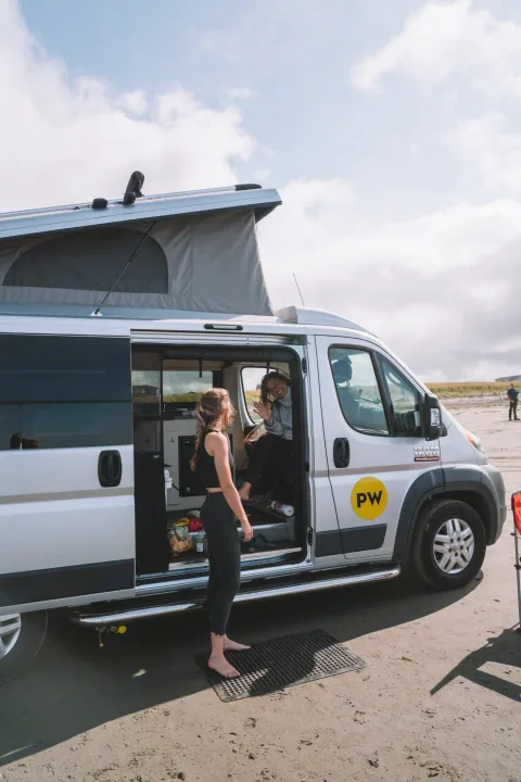 Two women by their pop-top rv rental camper van on a sandy spot. One stands barefoot outside, another waves from inside.. Ram Pleasure Way 2019