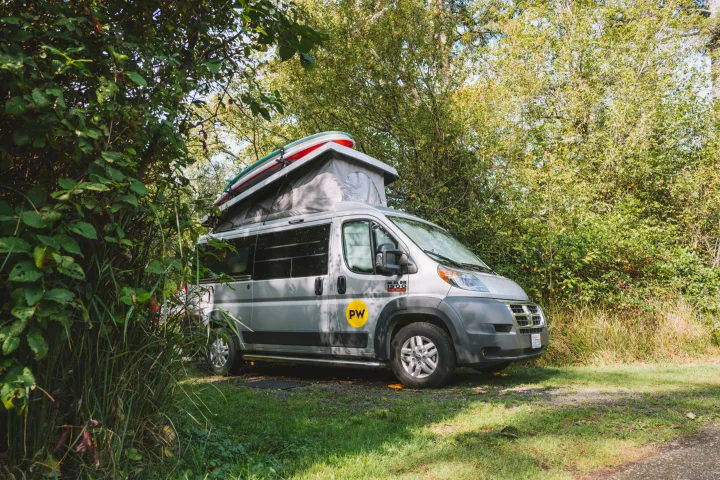 A grey camper van RV rental with a pop-top roof and paddleboards is parked amidst lush trees at a sunny campsite.. Ram Pleasure Way 2019