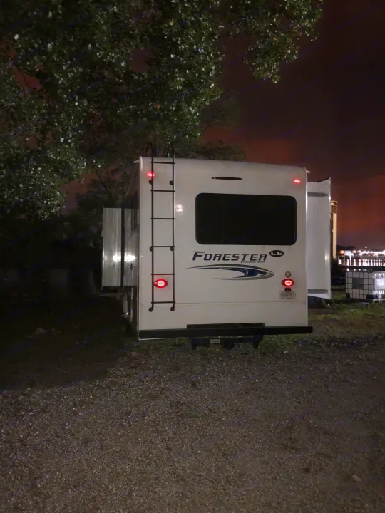 Rear view of a white Forester RV rental parked on gravel at night, with slide-outs extended and tail lights on.. Forest River forester 2019