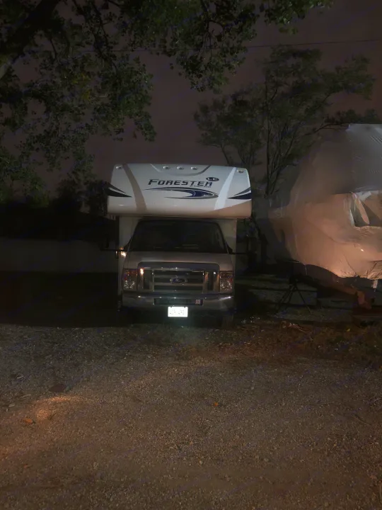 White Ford Forester RV rental brightly lit, parked on a gravel lot at night next to a tarp-covered vehicle.. Forest River forester 2019