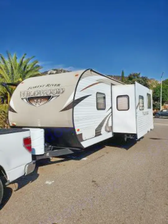 White Forest River Wildwood travel trailer RV rental, with slide-out, hitched to a truck on asphalt. Sunny day.. Forest River Wildwood 2016