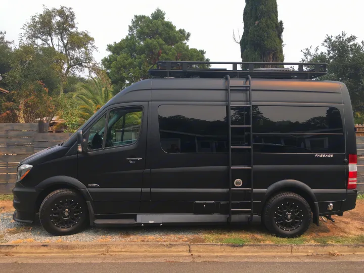 This sleek black Sprinter camper van, ideal for RV rental, features a roof rack, ladder, and rugged all-terrain tires.. Mercedes-Benz Sprinter 2018