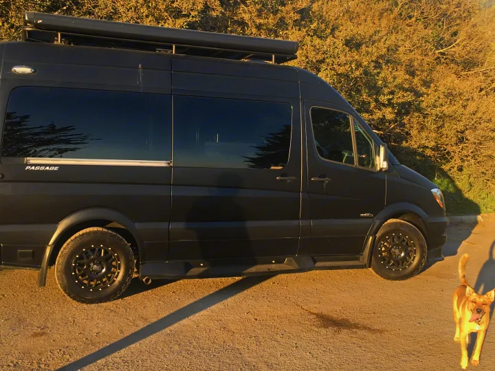 Black Midwest Passage camper van, great for rv rental, on dirt with a dog. Features off-road tires and roof rack.. Mercedes-Benz Sprinter 2018