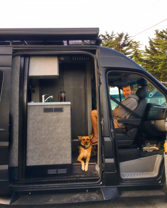 Man in driver's seat and dog in doorway of a dark RV rental van. Shows cozy interior kitchen. Perfect for pet-friendly adventures!. Mercedes-Benz Sprinter 2018