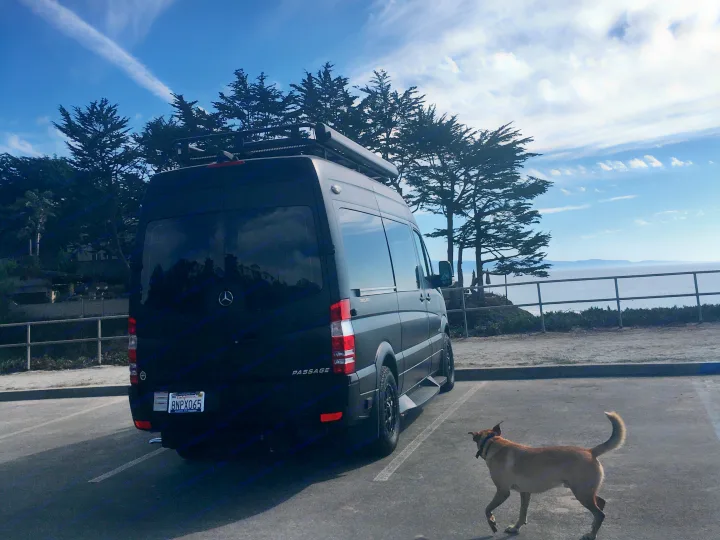 Black Mercedes Sprinter RV rental parked oceanside, with a dog exploring nearby under a blue sky.. Mercedes-Benz Sprinter 2018