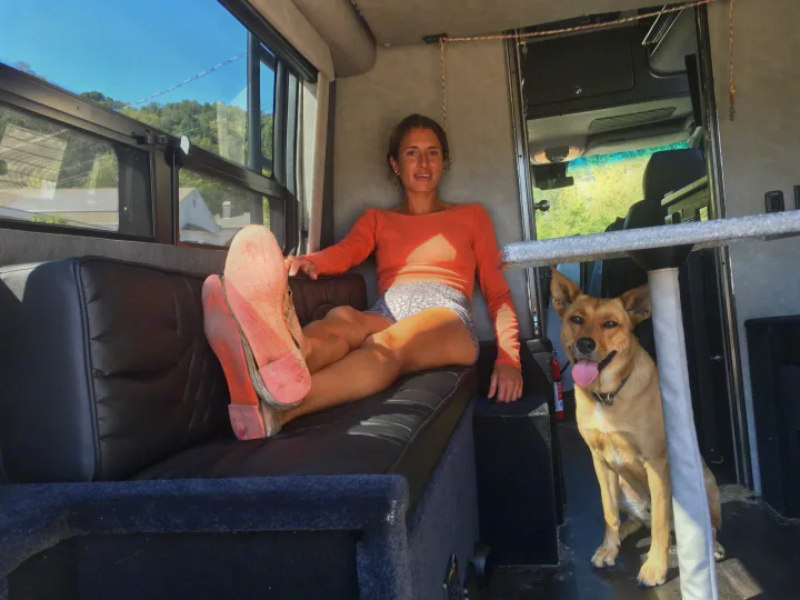 An RV rental interior: a woman relaxes on a bench with her feet up, and a smiling dog with its tongue out nearby.. Mercedes-Benz Sprinter 2018