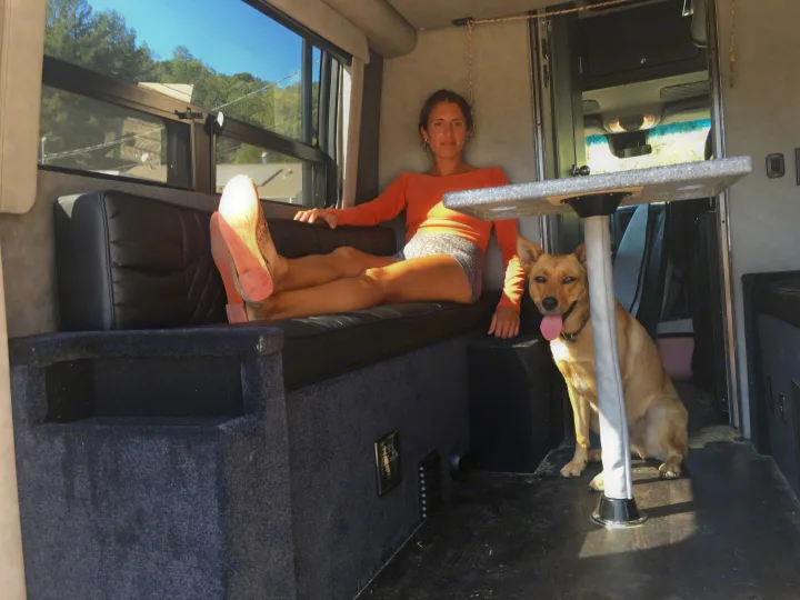 A woman and her dog relax inside a bright RV rental. She's on a black bench, feet up, next to a table. The tan dog sits nearby.. Mercedes-Benz Sprinter 2018