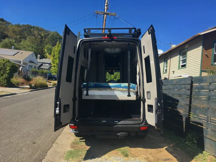 Rear view of an RV rental with open doors, showing a comfortable raised bed and ample storage. Ready for adventure!. Mercedes-Benz Sprinter 2018