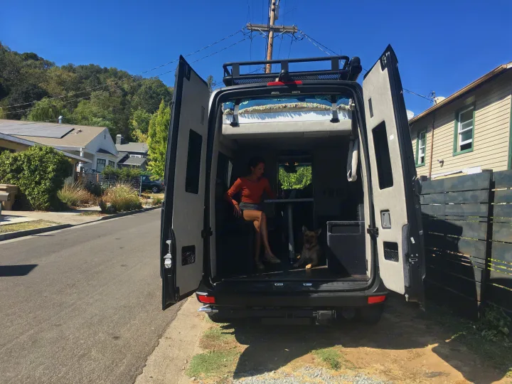 A woman and dog relax inside an rv rental camper van with its back doors open, parked on a sunny street.. Mercedes-Benz Sprinter 2018