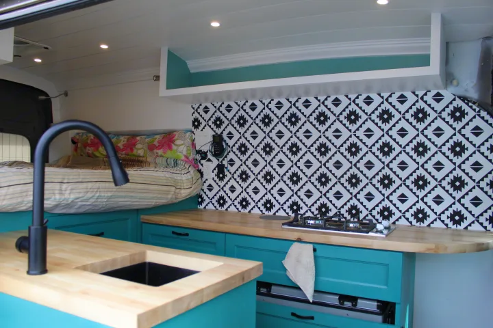 This RV rental interior showcases a cozy custom kitchenette with teal cabinets, wood counters, and a stylish backsplash, next to a raised bed.. Ford Transit 2020
