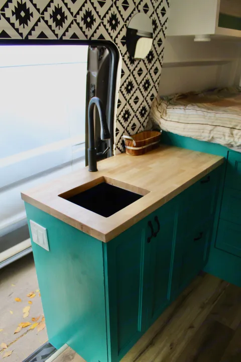 Custom RV rental interior: teal kitchen, wood counter, black sink, and a cozy bed. Leaves visible outside van door.. Ford Transit 2020