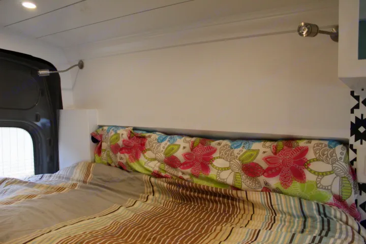 Inside an RV rental: a cozy bed with striped comforter, floral pillow, and two wall-mounted reading lights.. Ford Transit 2020
