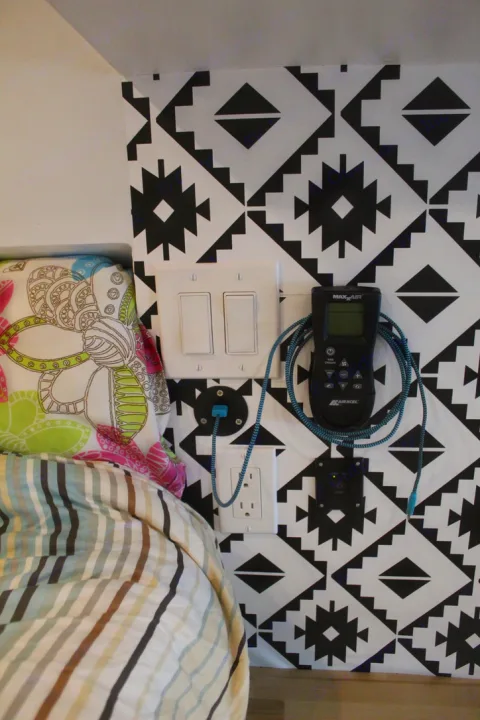 Convenient electrical setup in an rv rental, with outlets, light switches, and climate control remote on a patterned wall beside a bed.. Ford Transit 2020