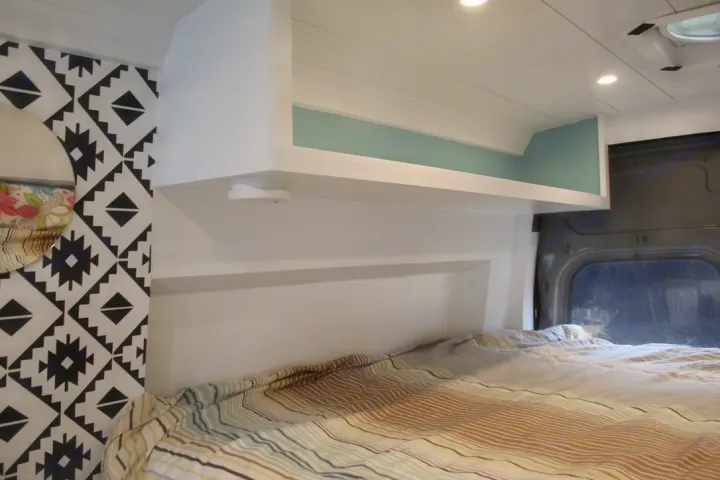 Interior of an RV rental featuring a cozy bed, geometric wall pattern, and integrated overhead storage with accent lighting.. Ford Transit 2020