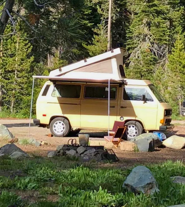 Tan pop-top camper van parked at a forest campsite with an awning extended, ideal for an rv rental adventure.. Volkswagen Westfalia 1982