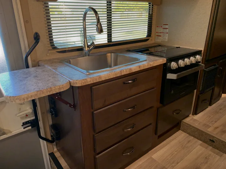 A functional RV rental kitchen with a stainless steel sink, pull-out faucet, stove/oven, dark cabinets, and extension counter.. Thor Freedom Elite 2021