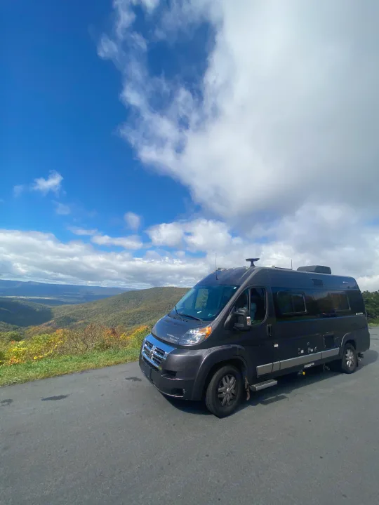 Dark grey Winnebago RV rental parked on a mountain road, offering stunning views of valleys & autumn foliage under a blue sky.. Winnebago Travato 2018
