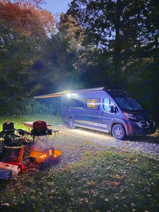 Cozy night camping with an RV rental: warm campfire, chairs, and an illuminated camper setup under the trees.. Winnebago Travato 2018