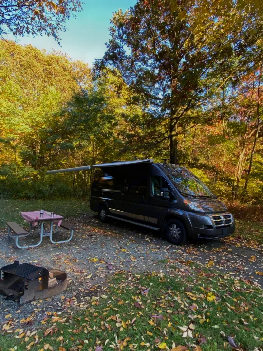 Experience autumn beauty in an RV rental! This camper van is set up at a vibrant fall campsite with a picnic table.. Winnebago Travato 2018