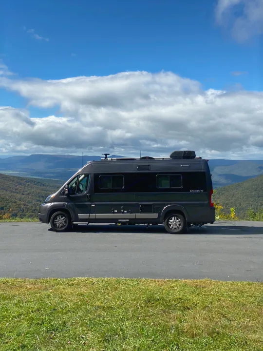 Black RV rental camper van parked on a paved mountain overlook, showcasing a vast landscape under cloudy skies.. Winnebago Travato 2018