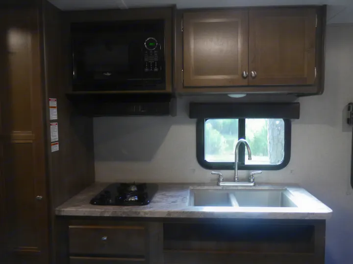This RV rental kitchen features a microwave, two-burner stovetop, double sink, and wooden cabinets, with a window looking out to trees.. Coleman Other 2018