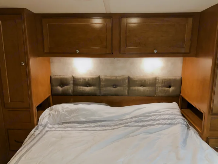 Cozy RV bedroom with a comfy bed, upholstered headboard, and wood storage. Perfect for your next RV rental adventure.. Winnebago Vista 2018