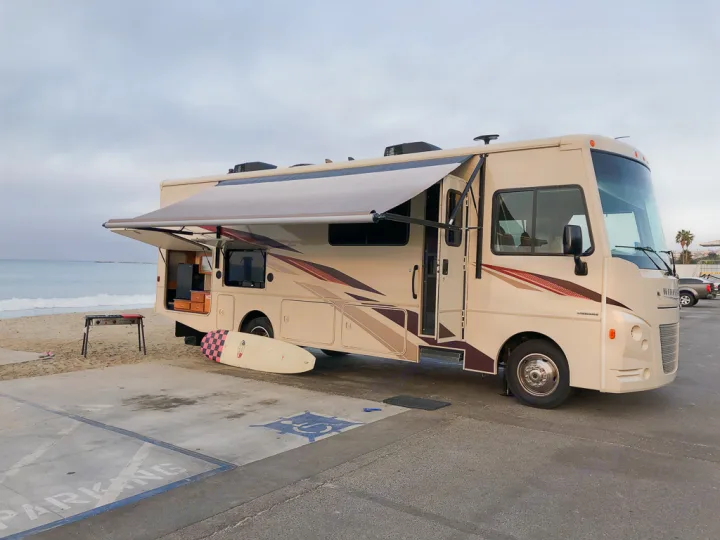Enjoying a beach day with an RV rental! This cream motorhome is parked oceanside with an awning out, surfboard, and grill.. Winnebago Vista 2018