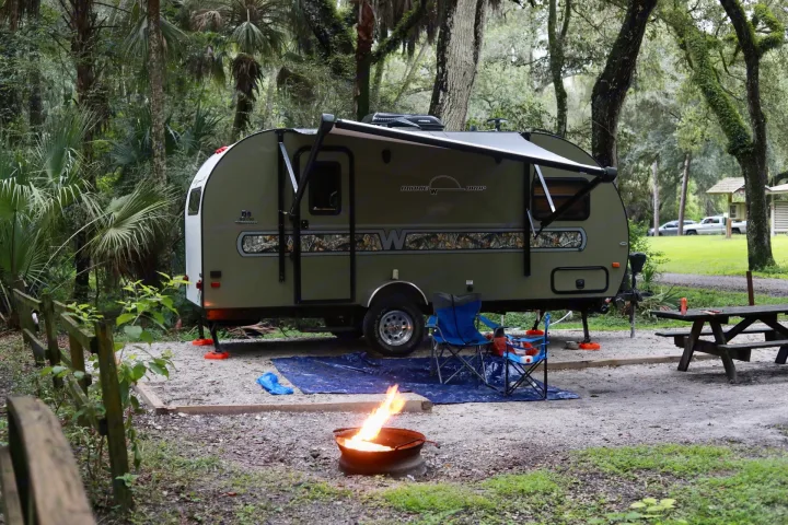 An olive green teardrop rv rental is set up on a gravel campsite under tall trees, with a bright campfire, chairs, and a picnic table.. Other Other 2018
