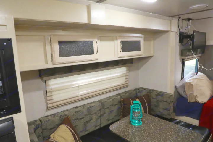Inside an RV rental: a comfortable dinette with a table & lantern, overhead cabinets, window blinds, and a bed area.. Other Other 2018