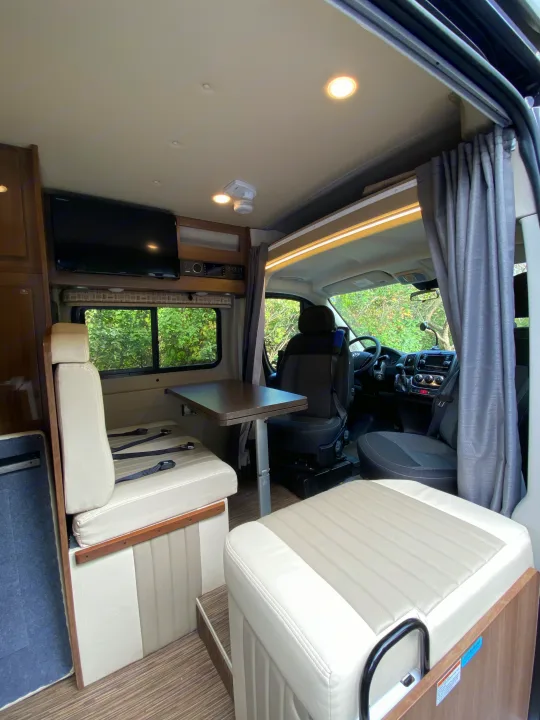 Inside a modern RV rental: dining table, comfy seats, TV, and driver's cabin view for your road trip comfort.. Winnebago Travato 2018