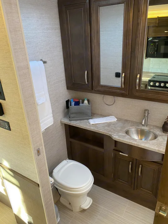 Clean RV rental bathroom with a toilet, vanity, and sink. A modern kitchen area is reflected in the mirror.. Entegra Coach Other 2021