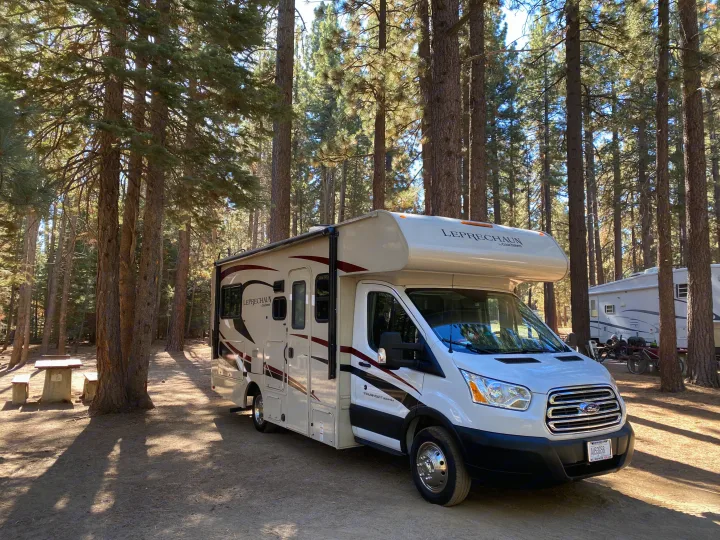 White RV rental motorhome parked in a sunny forest campsite, surrounded by tall pine trees and natural shadows.. Other Other 2020