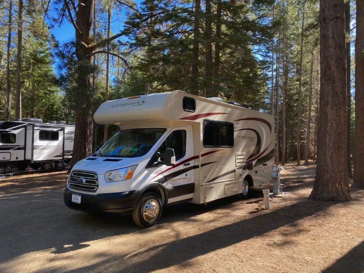A beige and white Class C RV rental is parked at a sunny forest campsite, surrounded by tall pine trees.. Other Other 2020