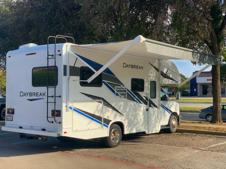 White "Daybreak" RV rental with awning extended, parked near trees and a building.. Thor Daybreak 2020
