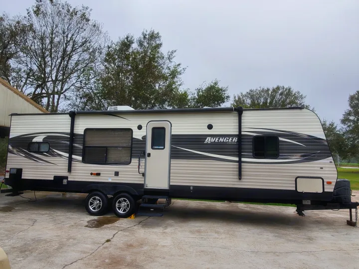 A cream and black travel trailer, perfect for your next rv rental, is parked on concrete with trees and overcast. Prime Time Avenger 2016