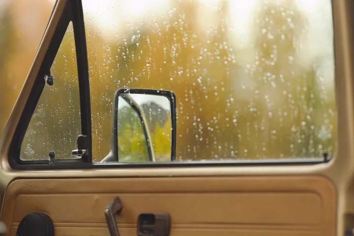 Inside an RV rental on a rainy autumn day. Raindrops on window, blurred fall trees, and a side mirror visible.. Volkswagen Westfalia 1986