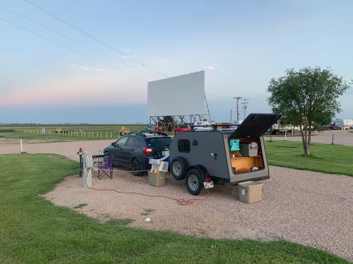 RV rental setup at a drive-in movie theater campsite. Subaru and teardrop trailer with open galley under dusk sky.. Little Guy Other 2020