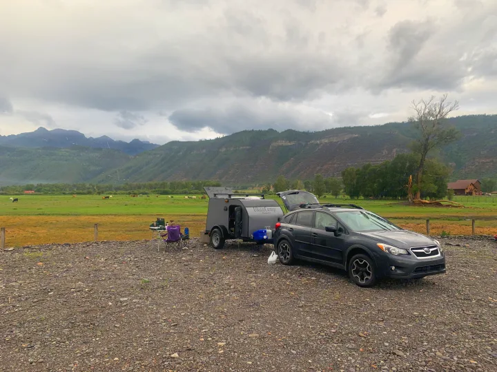 RV rental teardrop camper hitched to a dark SUV on a gravel lot. Scenic mountain and field view with grazing cows.. Little Guy Other 2020