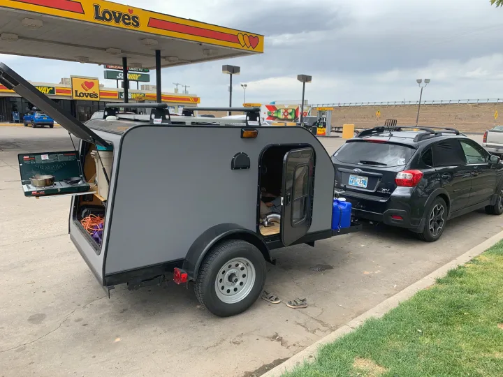 Compact teardrop RV rental trailer hitched to a Subaru at a Love's gas station, showing open rear kitchen and side door.. Little Guy Other 2020