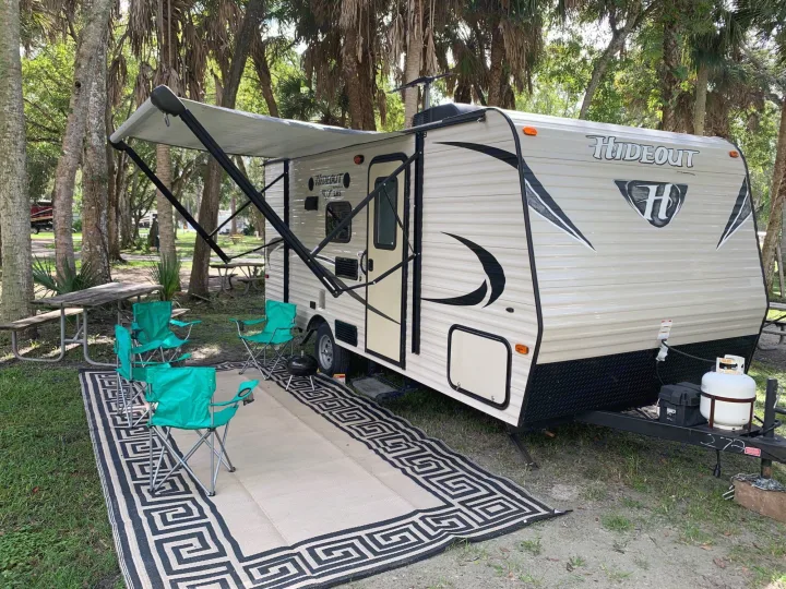 An RV rental travel trailer at a shaded campsite, featuring an extended awning, an outdoor rug, and four green chairs.. Keystone Hideout 2016