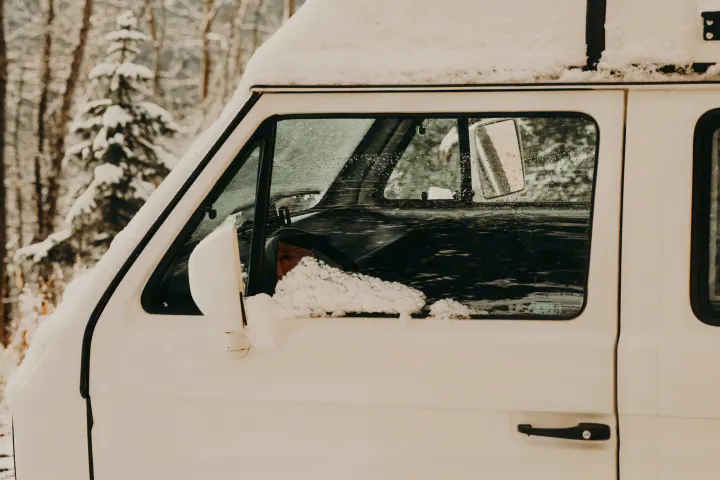 A white RV rental, covered in fresh snow, is parked in a blurry winter forest. Side view shows driver's window and mirror.. Volkswagen Weekender 1991