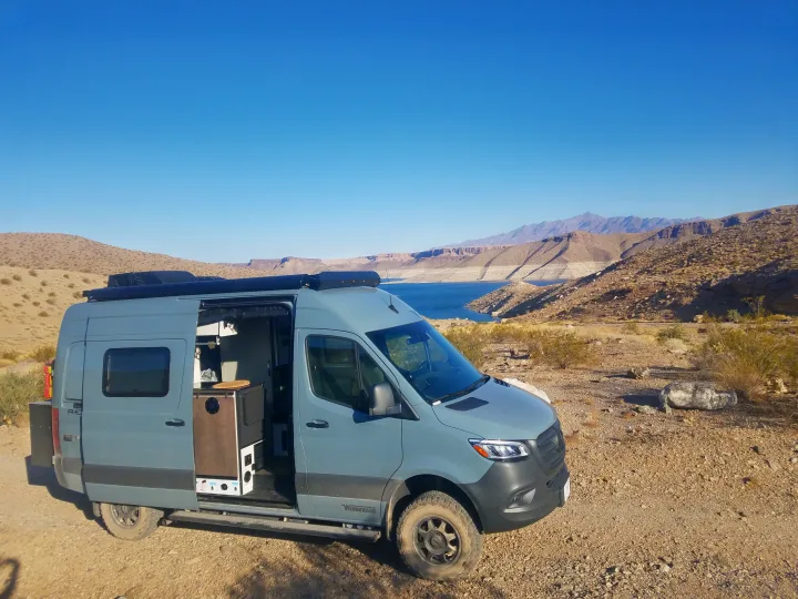 This grey RV rental is parked with its side door open, revealing its interior by a beautiful blue lake and arid mountains.. Winnebago Revel 2020