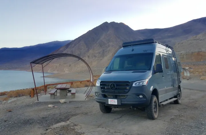 Gray Mercedes Sprinter rv rental on a scenic lake shore with mountains and a picnic table. Ideal for adventure.. Winnebago Revel 2020